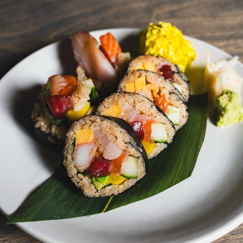 A white plate holds colorful sushi rolls, sashimi, yellow pickled ginger, and wasabi on a banana leaf.